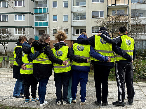 Gruppe von sieben Personen in gelben Warnwesten steht im Kreis und legt die Arme umeinander in einem Wohngebiet.