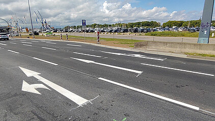 Mehrspurige Straße mit weißen Fahrbahnmarkierungen und einem nach links zeigenden Pfeil auf dem linken Fahrstreifen