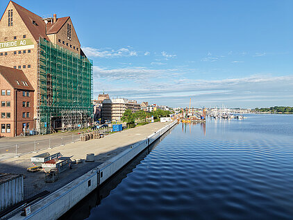 Blick auf einen Hafen mit Lagergebäuden, einem langen Kai und ruhigem Wasser unter blauem Himmel