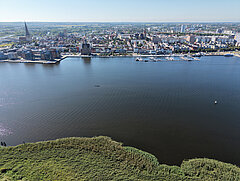 Luftaufnahme einer Stadt mit Fluss, im Vordergrund Schilf am Ufer und im Fluss mehrere kleine Boote