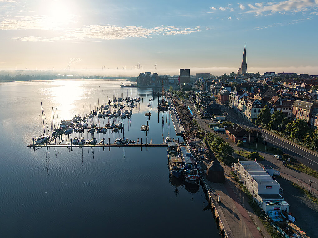 Luftaufnahme eines Stadthafens mit Segelbooten, Uferpromenade und Stadt mit Kirchturm im Hintergrund