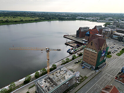 Blick auf einen Hafen mit einem großen Gebäude mit der Aufschrift 'GETREIDE AG', einem Baukran und einem im Bau befindlichen Gebäude am Wasser
