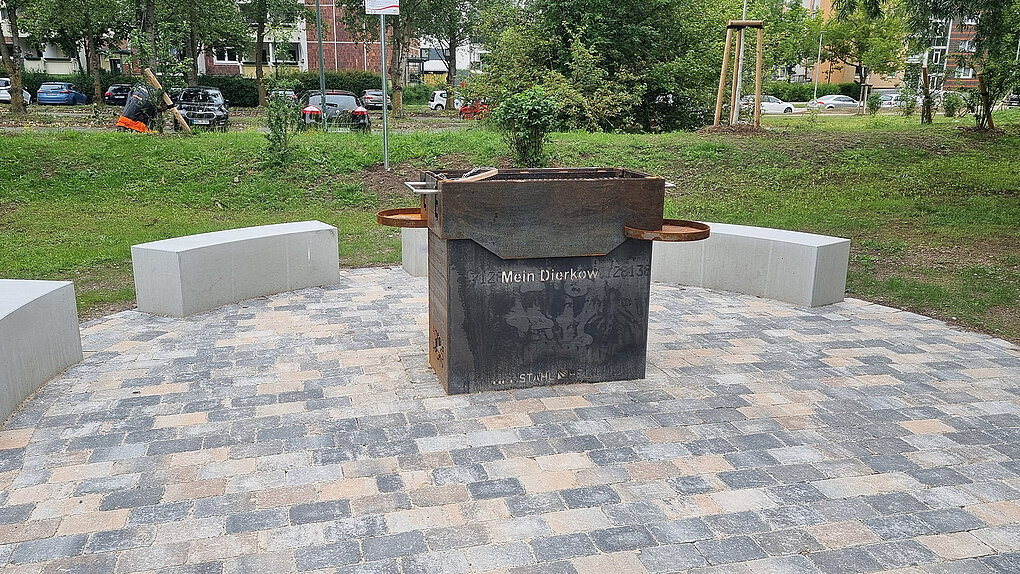 Runder Sitzbereich mit drei halbkreisförmigen Betonbänken um einen quadratischen Metallgrill auf gepflastertem Boden in einem Park