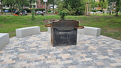 Runder Sitzbereich mit drei halbkreisförmigen Betonbänken um einen quadratischen Metallgrill auf gepflastertem Boden in einem Park