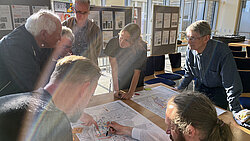 Sieben Personen diskutieren konzentriert über Pläne auf einem Tisch in einem hellen Raum mit großen Fenstern und Informationswänden im Hintergrund.