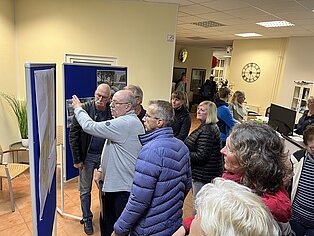 Gruppe von Menschen in einem Raum, die an einer Informationswand stehen und diskutieren. Einige Personen zeigen auf den Inhalt der Wand, während andere aufmerksam zuhören.