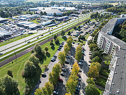 Luftaufnahme eines Parkplatzes neben einer Straße mit mehreren parkenden Autos und angrenzenden Gebäuden in einer grünen Landschaft