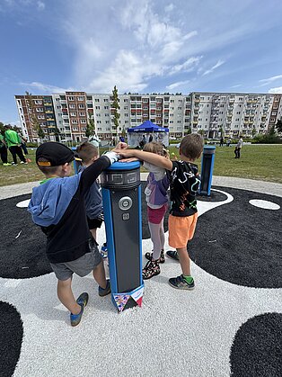 VierKinder bedienen gemeinsam eine blaue Säule auf einem Spielplatz mit Kiesflächen und Wohngebäuden im Hintergrund