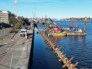 Bauarbeiten am Stadthafen mit Schwimmbagger und Baugerüst im Wasser, Arbeiter und Fahrzeuge am Ufer bei klarem Himmel