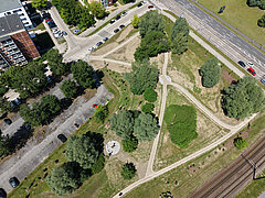 Luftaufnahme eines dreieckigen Parks mit Wegen, Bäumen, einem kleinen runden Sitzbereich und angrenzenden Straßen sowie Bahngleisen
