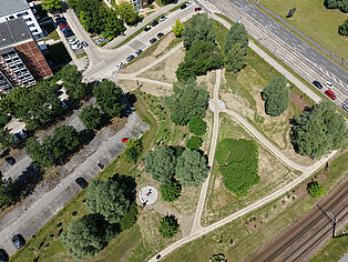 Luftaufnahme eines dreieckigen Parks mit Wegen, Bäumen, einem kleinen runden Sitzbereich und angrenzenden Straßen sowie Bahngleisen