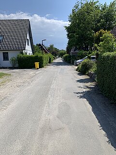 Kaputte Straße zwischen Häusern mit Bäumen und Sträuchern an der Seite unter bewölktem Himmel.