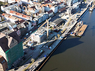 Luftaufnahme eines Stadthafens mit Baustelle, Kran, parkenden Autos und angrenzenden Wohngebäuden am Flussufer