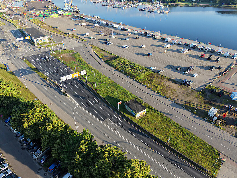 Luftaufnahme einer mehrspurigen Straße mit Rechtsabbiegerspur neben einem großen Parkplatz und Wasser im Hintergrund