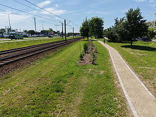 Blick auf einen schmalen Fußweg neben einer Bahnstrecke mit Oberleitungen und einer Straße mit Fahrzeugen im Hintergrund