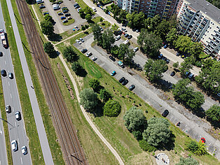 Luftaufnahme eines Wohngebiets mit mehrstöckigen Häusern, Parkplätzen, Bäumen, einer Straße mit Autos und parallel verlaufenden Bahngleisen