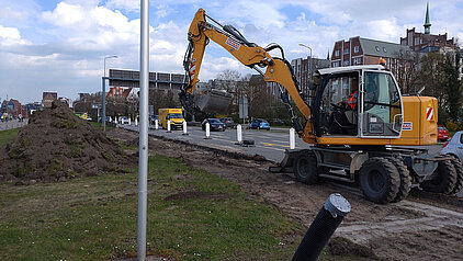 Bagger mit ausgefahrenem Arm auf einer Baustelle neben einer Straße mit mehreren Fahrzeugen im Hintergrund