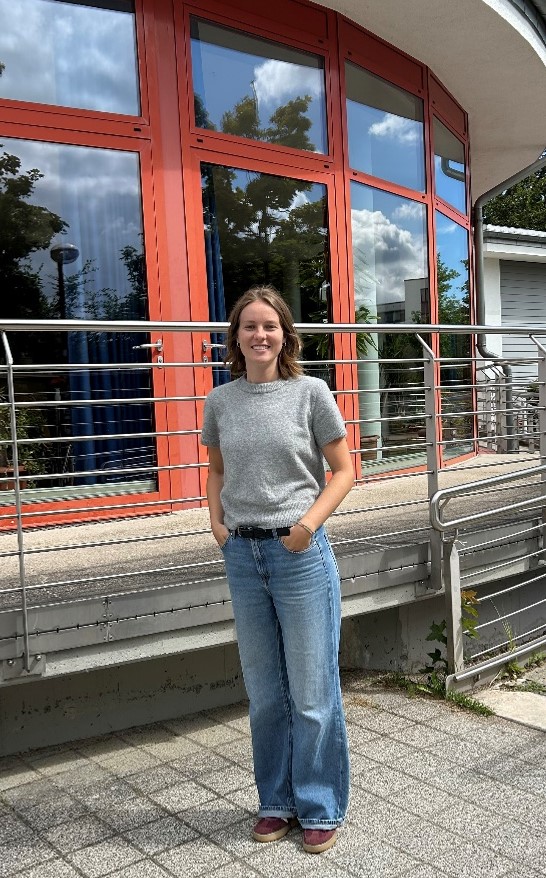 Frau im grauen T-Shirt und blauer Hose steht vor einem Fenster und lächelt in die Kamera.