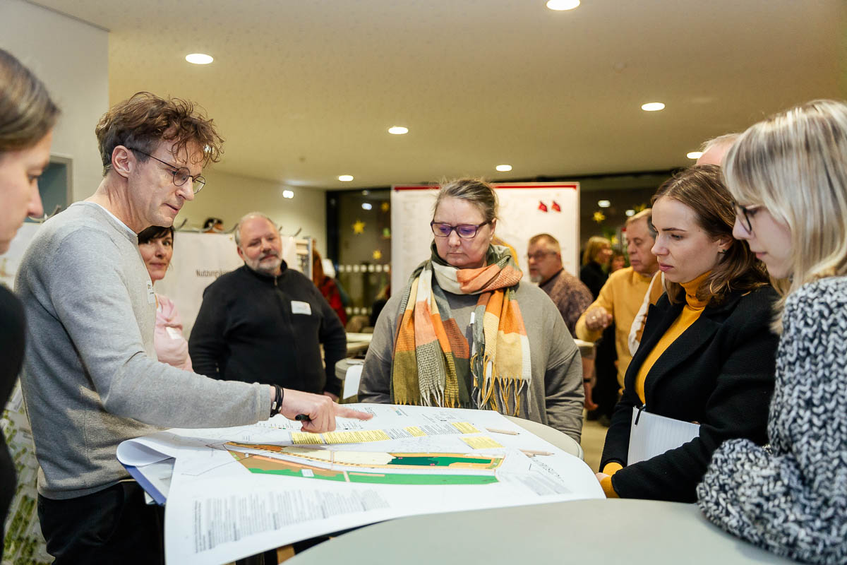 Gruppe von Menschen diskutiert um einen Tisch mit Plänen oder Karten in einem Innenraum.